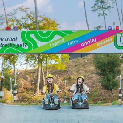 Two girl friends ride underneath a large banner showing off the different Luge tracks at Skyline Luge Tongyeong.
